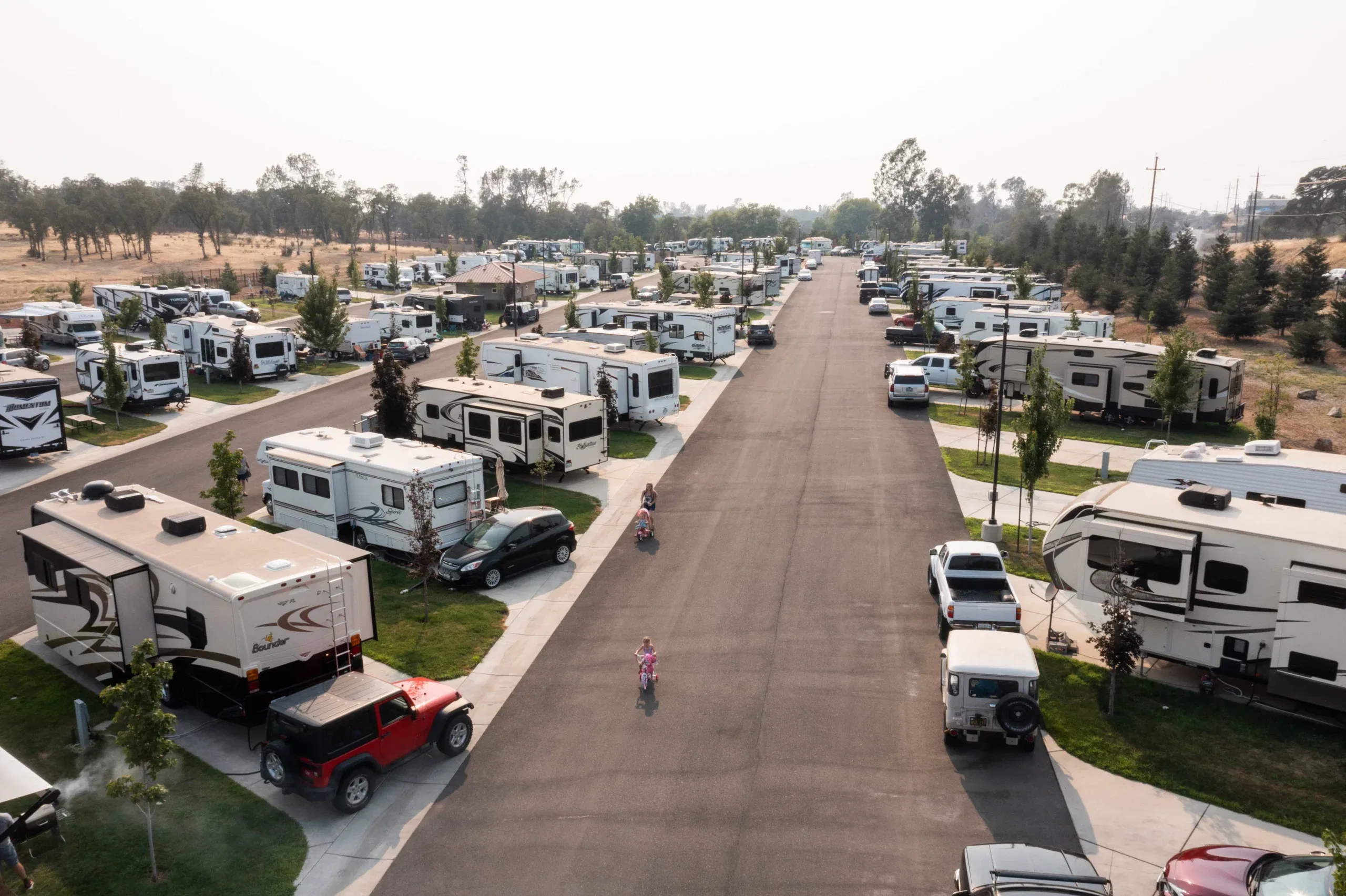 Aerial view of an RV park with numerous parked trailers, cars, and trucks along paved roads; two people ride bicycles down the main road.