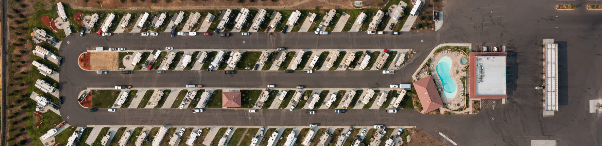 Aerial view of a neatly arranged RV park with rows of parked RVs, cars, a swimming pool, and a building on paved lots.