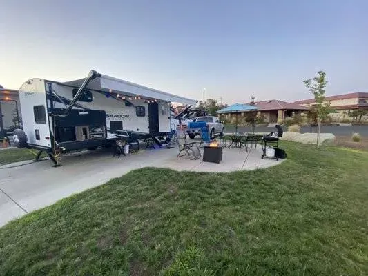 A travel trailer with an extended awning is parked on a driveway. A pickup truck, outdoor chairs, a table, and a fire pit are set up on a grassy area beside the trailer.