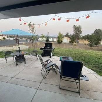 Patio with chairs, a table with an umbrella, a barbecue grill, and string lights, overlooking a grassy yard and trees.