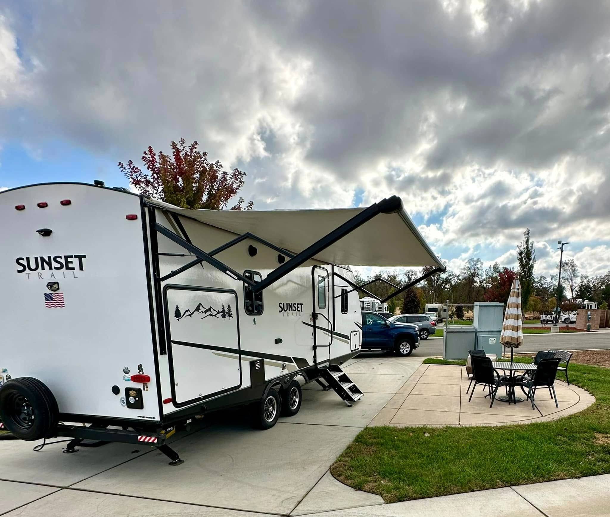 A white Sunset Trail travel trailer with an extended awning is parked on a concrete pad next to a patio set and umbrella under a cloudy sky.