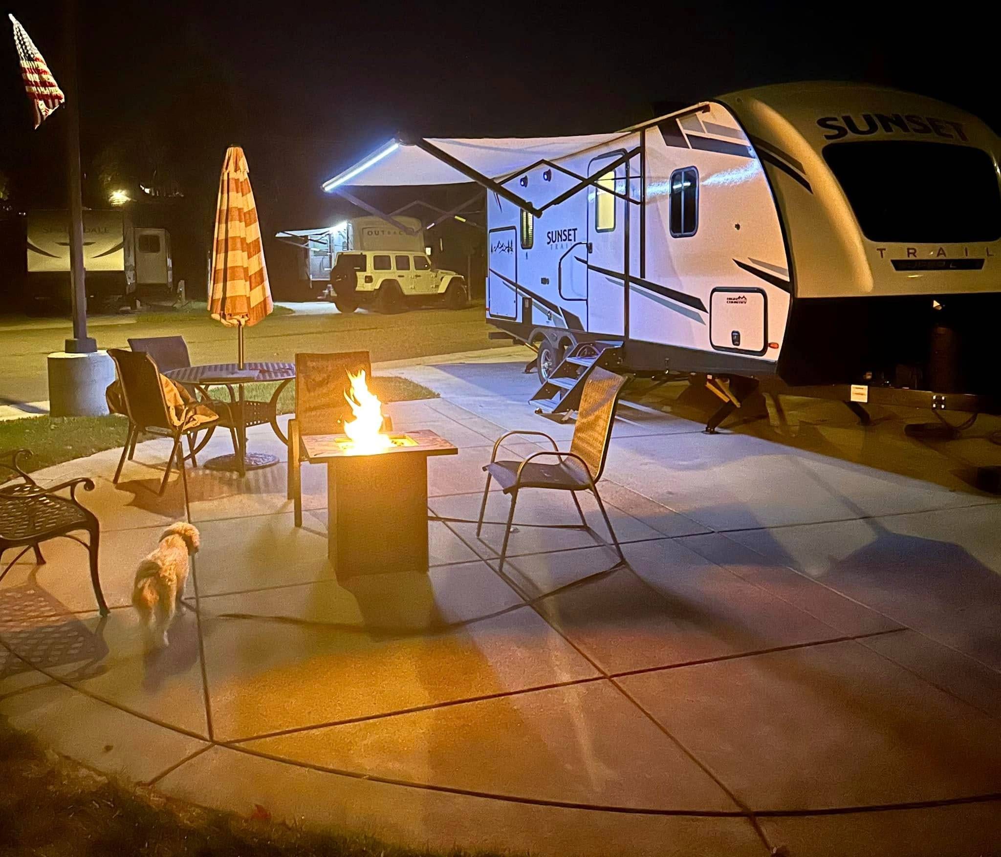 A lit fire pit with chairs sits on a patio beside a camper at night; a small dog walks nearby and an umbrella and American flag are visible.