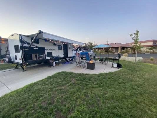 A travel trailer with an extended awning is parked on a paved area with outdoor seating, a fire pit, and a grill setup on the grass nearby.
