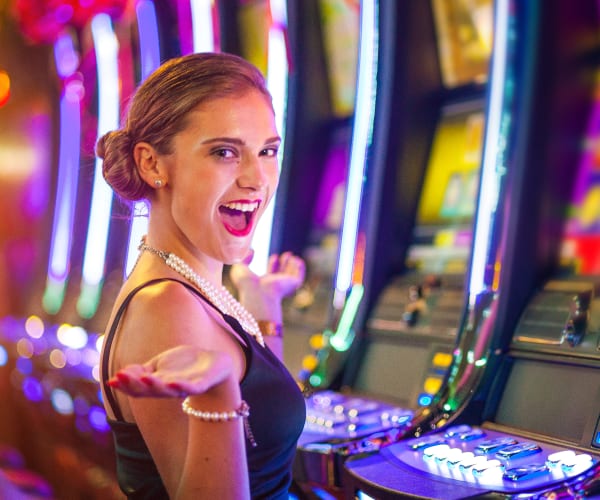 A woman in a black dress stands in front of brightly lit slot machines, smiling with her hands raised in a gesture of excitement.